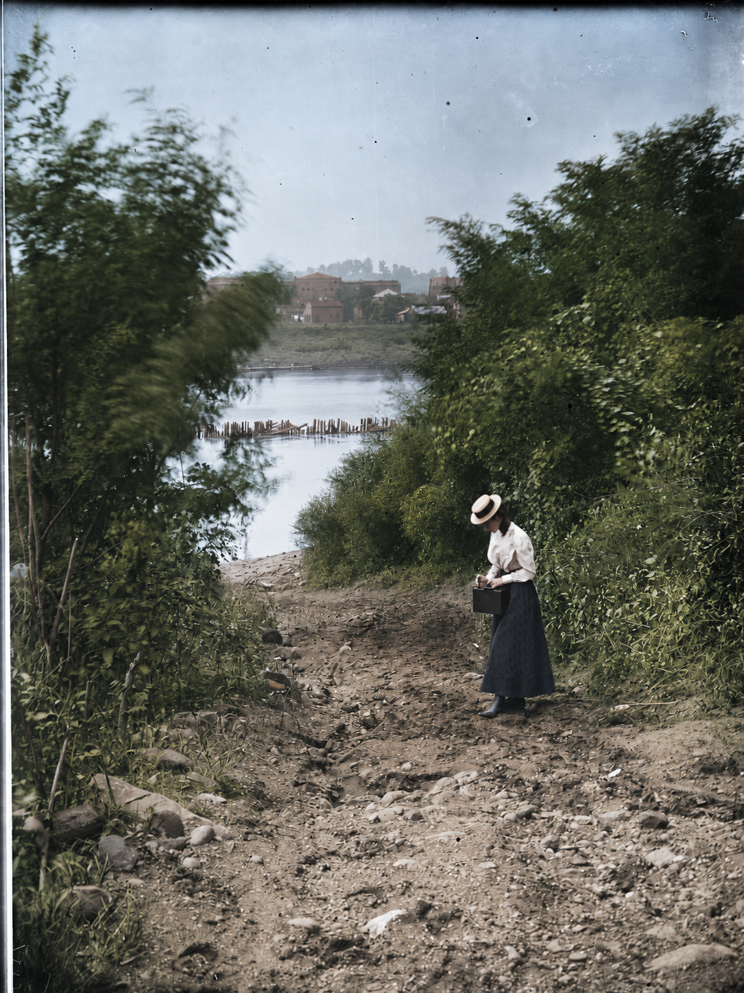 Colorized riverbank scene with a woman carrying a case